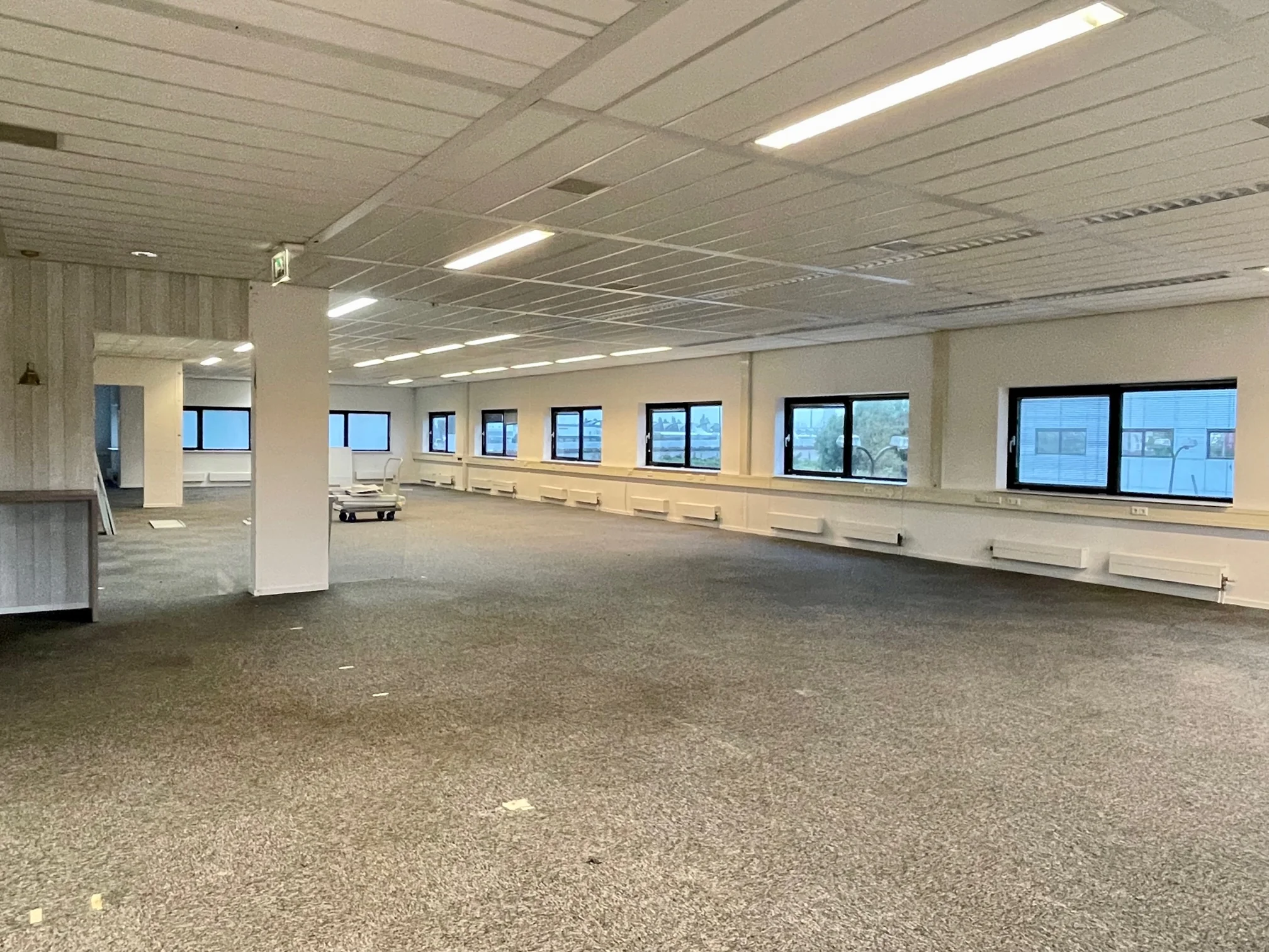 Lege kantoorruimte aan de Karel Doormanweg met grijze vloerbedekking en meerdere ramen langs de muur.